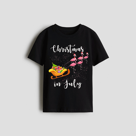 A black t-shirt with a Christmas-themed design featuring a sled with a Santa Claus figure and flamingos flying around it. The text on the t-shirt reads "Christmas in July".