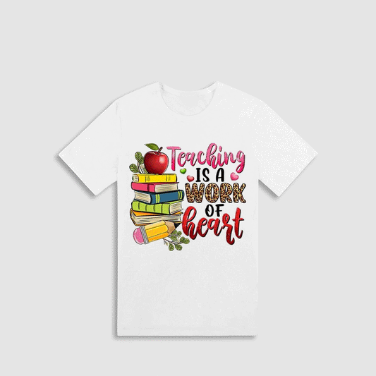 A white t-shirt with a colorful graphic design featuring a stack of books, an apple, and the text "Teaching is a Work of Heart".