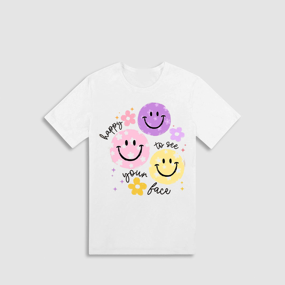 A white t-shirt with a graphic design featuring three smiling faces in different colors - pink, purple, and yellow - along with the text "hope to see your face" written around the edges.