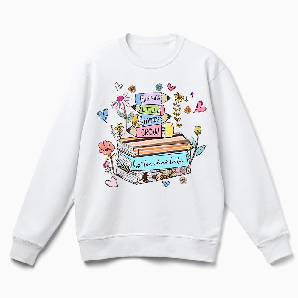 A white sweatshirt with a colorful graphic design featuring a stack of books, flowers, and hearts, along with the text "Helping little minds grow" and "Teacher's life".