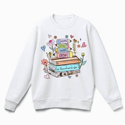 A white sweatshirt with a colorful graphic design featuring a stack of books, flowers, and hearts, along with the text "Helping little minds grow" and "Teacher's life".