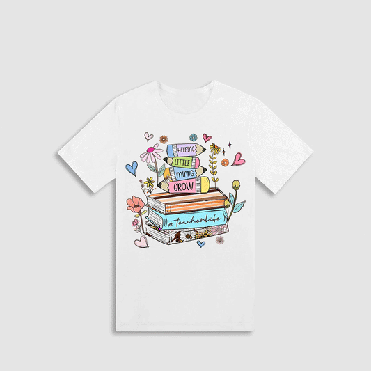A white t-shirt with a colorful graphic design featuring a stack of books, flowers, and other whimsical elements.